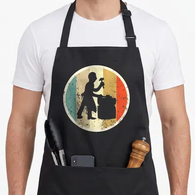Tablier de cuisine noir avec illustration circulaire d'un maréchal-ferrant forgeron en silhouette grise sur fond de bandes colorées délavées.
