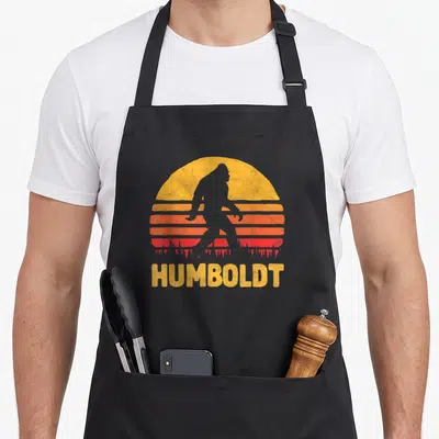 Tablier de cuisine noir avec silhouette de Sasquatch devant un soleil rayé et le mot Humboldt en jaune.