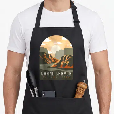 Tablier de cuisine noir avec une illustration archi-forme du Grand Canyon aux teintes orange et vert olive, surmontée du texte « GRAND CANYON NATIONAL PARK » en blanc et orange.