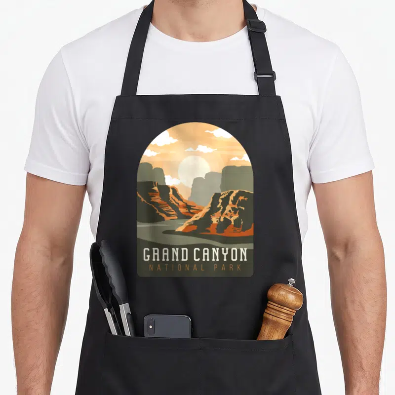 Tablier de cuisine noir avec une illustration archi-forme du Grand Canyon aux teintes orange et vert olive, surmontée du texte « GRAND CANYON NATIONAL PARK » en blanc et orange.