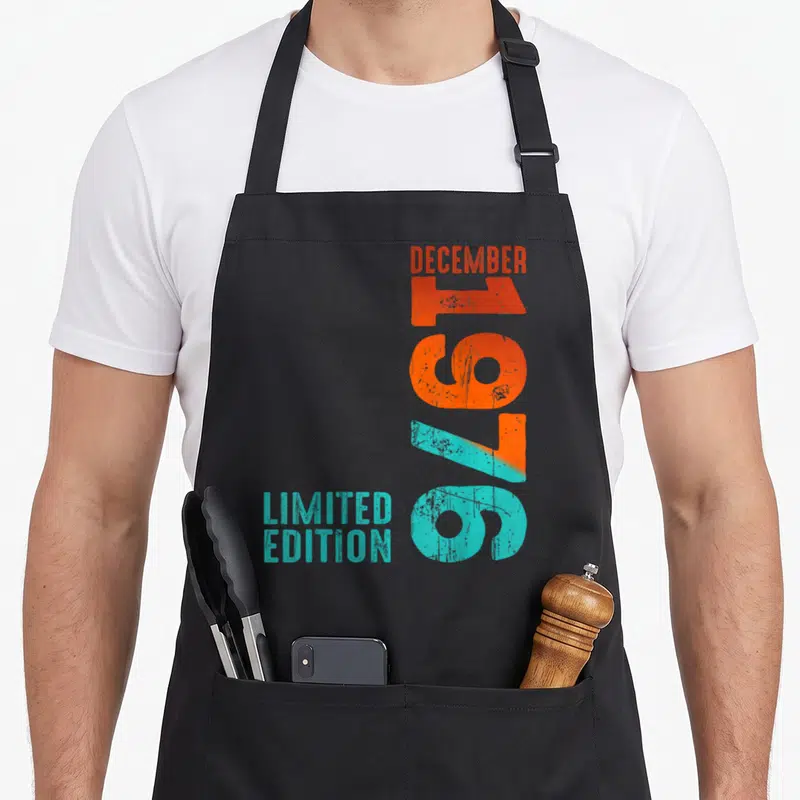 Tablier de cuisine noir avec l'inscription Décembre 1976 Limited Edition en lettres délavées orange et turquoise.