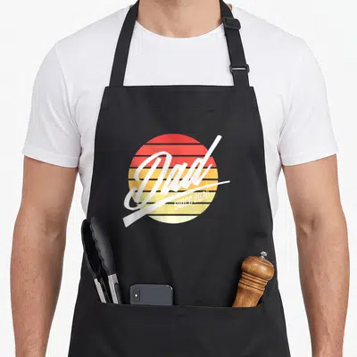 Tablier de cuisine noir avec logo rond Dad Since 2021 sur fond de soleil rétro orange et jaune.