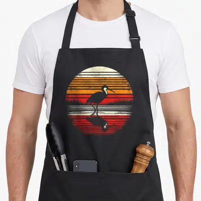 Tablier de cuisine noir avec illustration circulaire d'un oiseau jacana sur fond de soleil couchant orange et rouge style rétro.