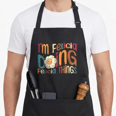 Tablier de cuisine noir avec typographie multicolore et fleur blanche au visage souriant entourée d'étoiles scintillantes.