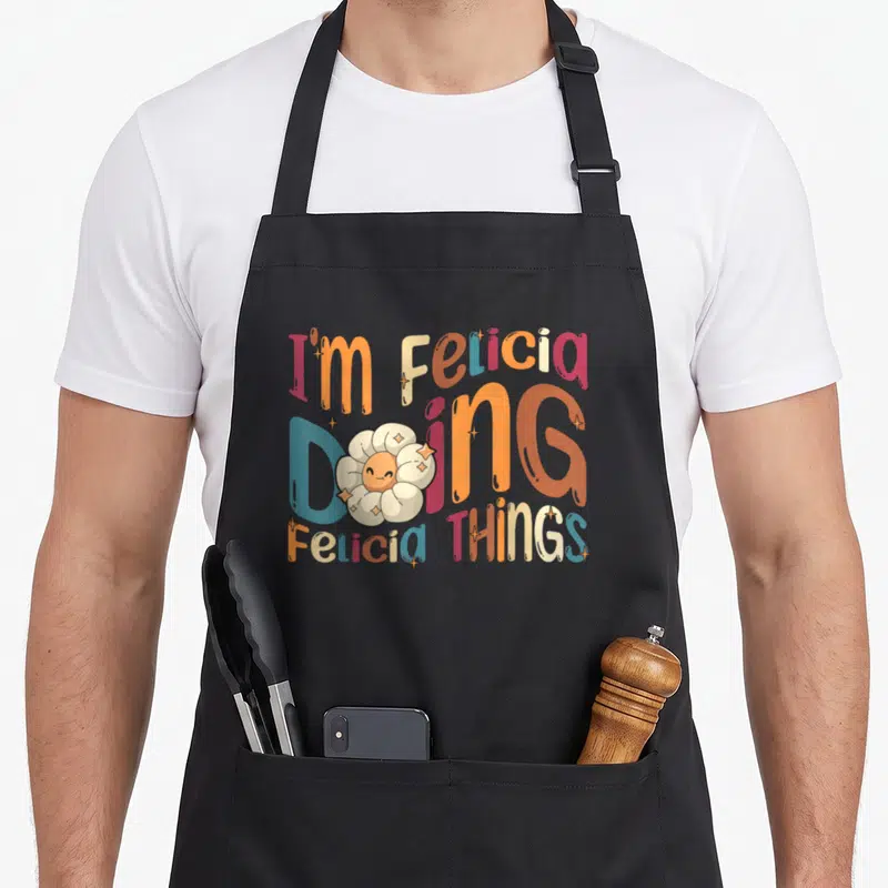 Tablier de cuisine noir avec typographie multicolore et fleur blanche au visage souriant entourée d'étoiles scintillantes.