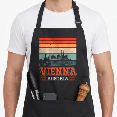 Tablier de cuisine noir avec illustration rétro du skyline de Vienne et texte Vienna Austria en orange et blanc.