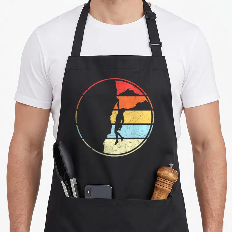 Tablier de cuisine noir avec illustration circulaire d'un grimpeur sur fond de soleil couchant multicolore.