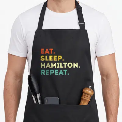 Tablier de cuisine noir avec le texte Eat Sleep Hamilton Repeat imprimé en lettres colorées style vintage.