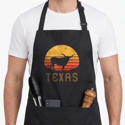 Tablier de cuisine noir avec silhouette de vache Longhorn et mot TEXAS sur fond de soleil orange rétro.