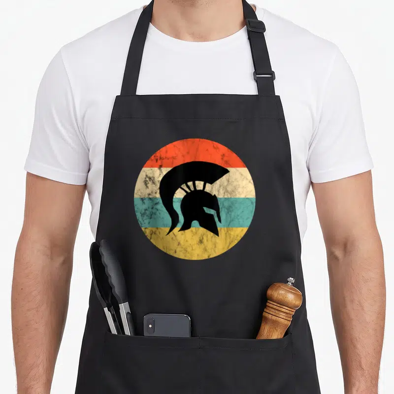 Tablier de cuisine noir avec illustration circulaire d'un casque gladiator spartano retro sur fond de coucher de soleil multicolore.