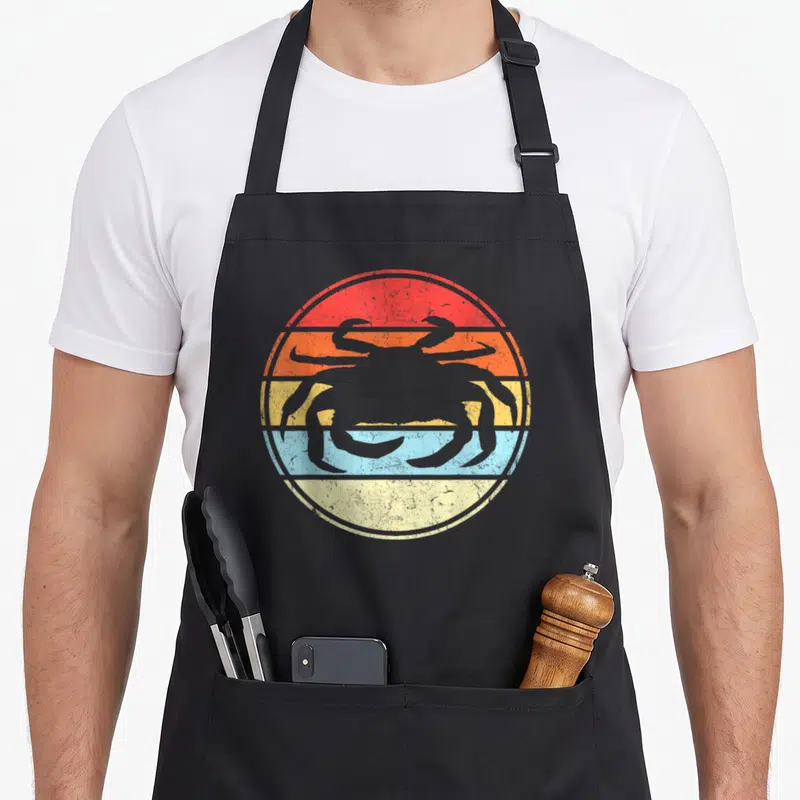 Tablier de cuisine noir avec une silhouette de crabe grise sur fond de cercle aux bandes colorées rouges, oranges, jaunes et bleues.