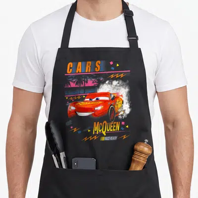 Tablier de cuisine noir avec Lightning McQueen rouge effectuant un dérapage sous un ciel rose et des palmiers stylisés.