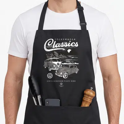 Tablier de cuisine noir avec illustration blanche d'une Beetle et d'un bus T1 devant des montagnes sous le texte Volkswagen Classics.