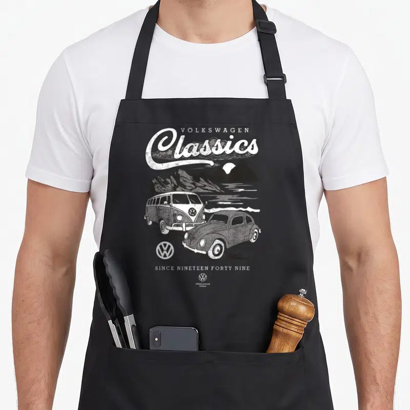 Tablier de cuisine noir avec illustration blanche d'une Beetle et d'un bus T1 devant des montagnes sous le texte Volkswagen Classics.
