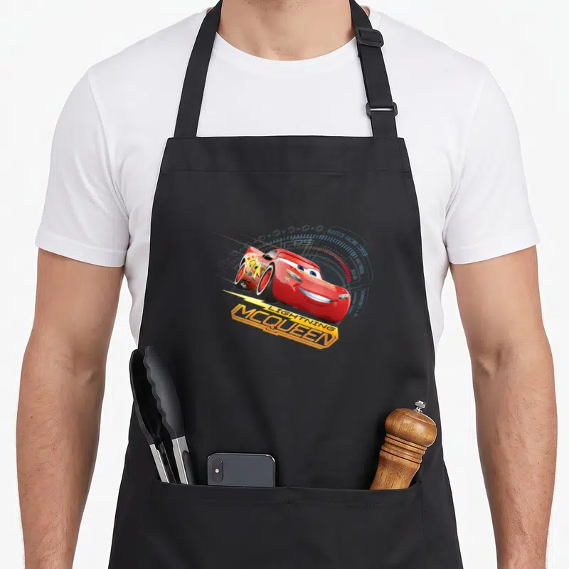 Tablier de cuisine noir avec Lightning McQueen rouge, le chiffre 95 et l'inscription McQueen en lettres orange dynamiques sur fond de compteur.
