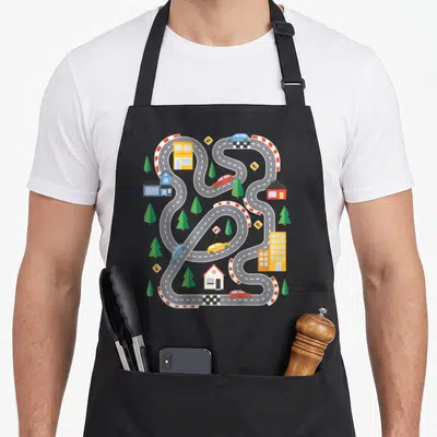 Tablier de cuisine noir avec un circuit de course en forme de route sinueuse, des voitures et des bâtiments colorés.