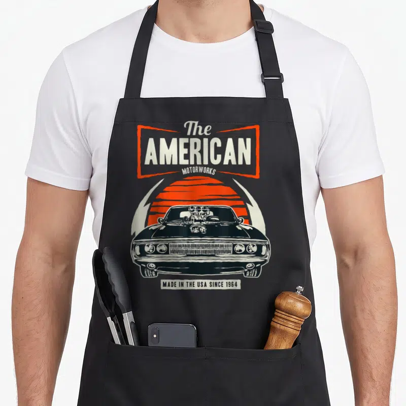 Tablier de cuisine noir avec une illustration de muscle car noire, un soleil orange rayé et le texte The American Motorworks en typographie vintage.