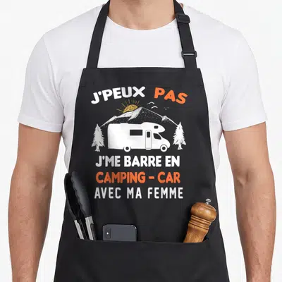 Tablier de cuisine noir avec illustration blanche d'un camping-car, sapins et soleil, surmonté du texte J'peux pas j'me barre en camping-car avec ma femme en blanc et orange.