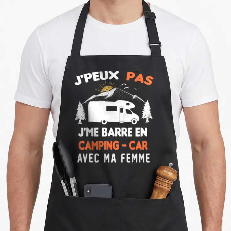 Tablier de cuisine noir avec illustration blanche d'un camping-car, sapins et soleil, surmonté du texte J'peux pas j'me barre en camping-car avec ma femme en blanc et orange.