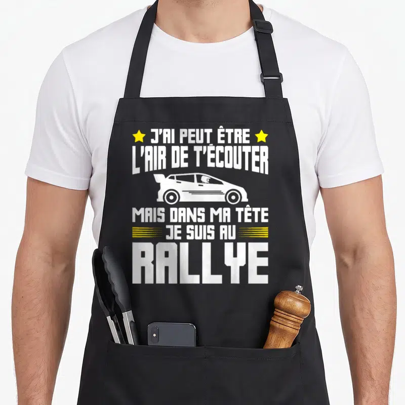 Tablier de cuisine noir avec une illustration de voiture blanche et le texte J'ai peut être l air de t écouter mais dans ma tête je suis au rallye en blanc et jaune.