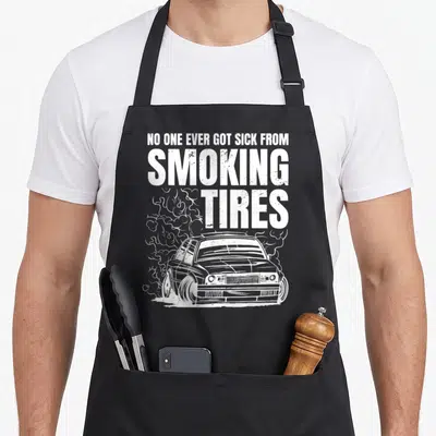 Tablier de cuisine noir avec illustration blanche d'une voiture en drift entourée de fumée et texte Smoking Tires.