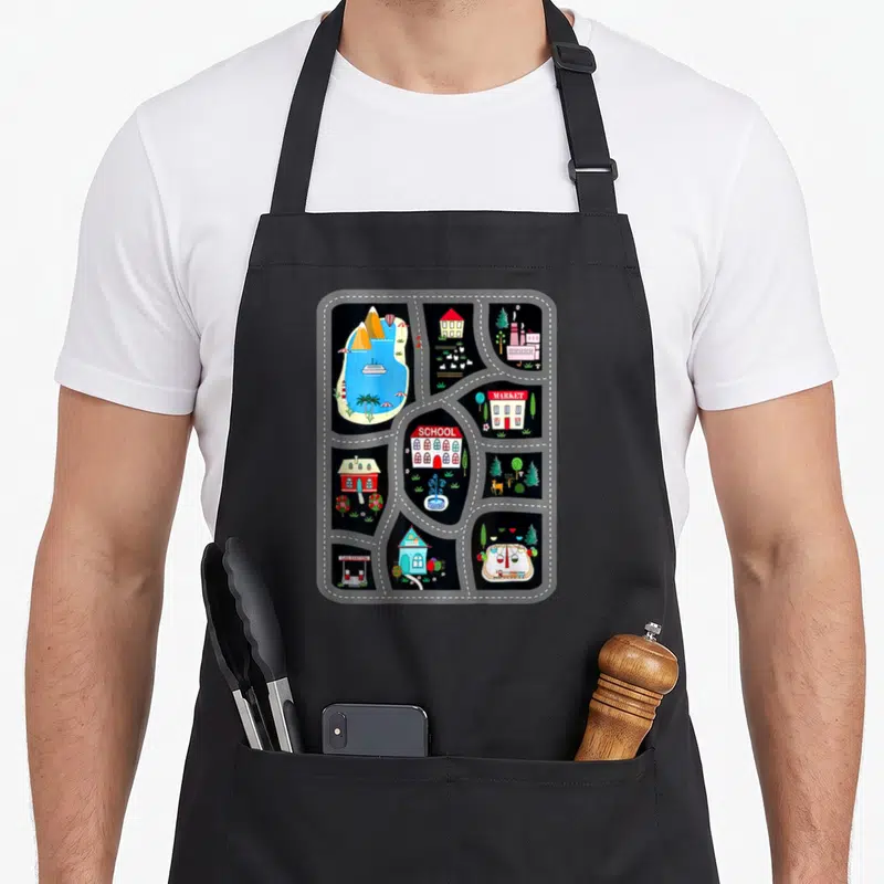 Tablier de cuisine noir avec motif carte de jeu routière colorée incluant école, marché et paysage.