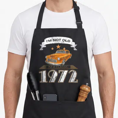 Tablier de cuisine noir avec une automobile orange vintage, des étoiles oranges et le texte blanc « I'M NOT OLD I'M A CLASSIC 1972 » sur fond texturé.
