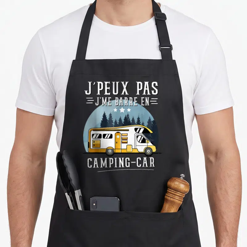 Tablier de cuisine noir avec illustration de camping-car jaune et texte blanc sur fond de forêt.