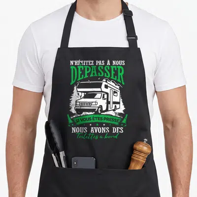 Tablier de cuisine noir avec illustration de camping-car et texte vert blanc humoristique sur fond uni.