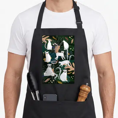 Tablier de cuisine noir avec une illustration de lémuriens blancs et de feuilles tropicales sur fond vert foncé.