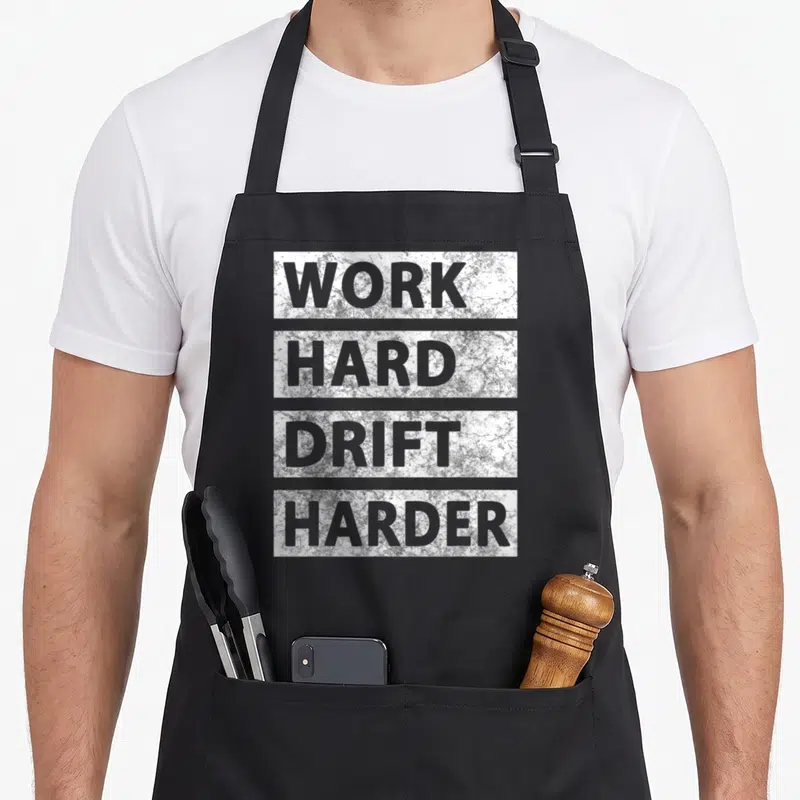 Tablier de cuisine noir avec inscription Work Hard Drift Harder en lettres capitales grises sur fond texturé blanc et gris.