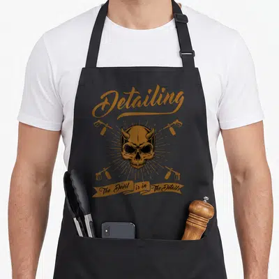 Tablier de cuisine noir avec illustration vintage d'un crâne cornu entouré de pistolets pulvérisateurs orange.