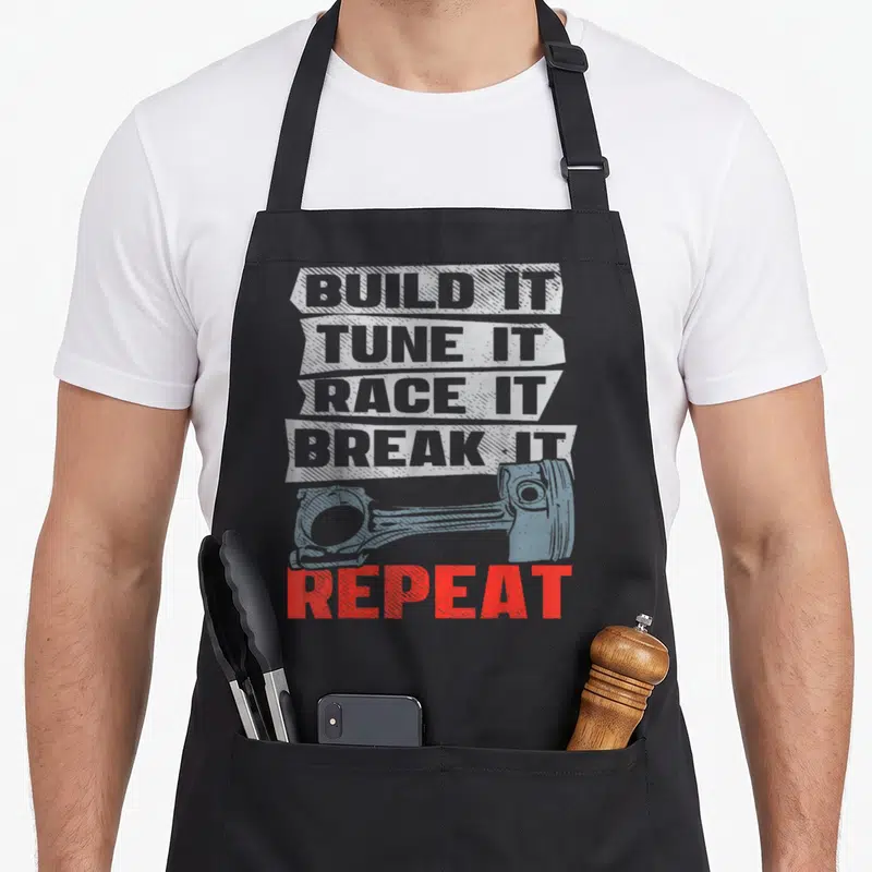 Tablier de cuisine noir avec le texte Build It Tune It Race It Break It Repeat et un piston bleu en arrière-plan.