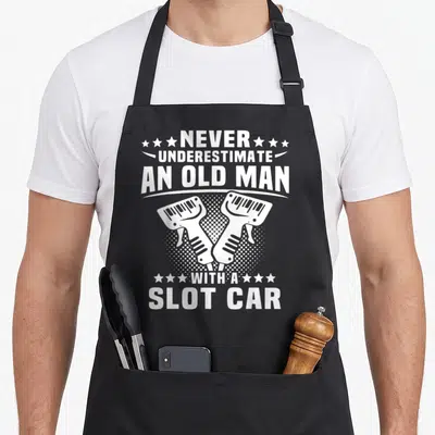 Tablier de cuisine noir avec texte blanc et illustration de deux manettes de slot car croisées.