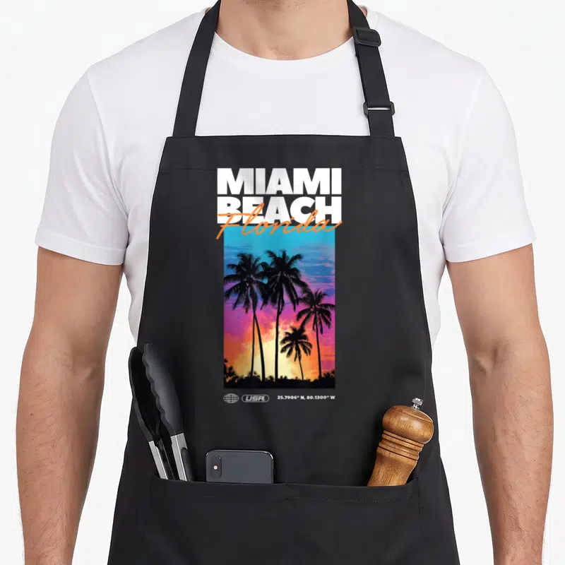 Tablier de cuisine noir avec illustration de palmiers au coucher de soleil et texte Miami Beach Florida USA.
