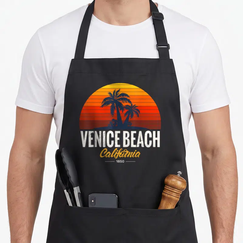 Tablier de cuisine noir avec illustration Venice Beach California 1850 et palmiers sur fond de soleil couchant dégradé.