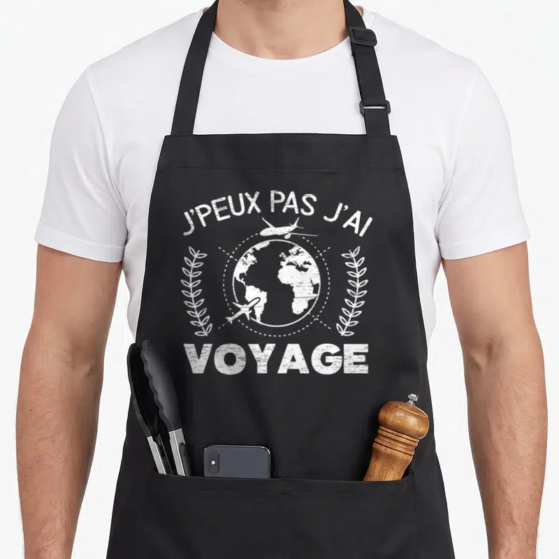 Tablier de cuisine noir avec inscription blanche J'peux pas j'ai voyage surmontant une illustration de globe terrestre et d'avions.