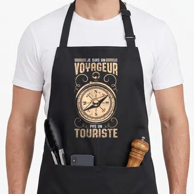 Tablier de cuisine noir avec une illustration de boussole vintage et le texte Je suis un voyageur pas un touriste imprimé en beige.
