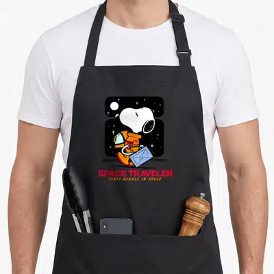 Tablier de cuisine noir avec Snoopy en astronaute orange portant une valise bleue sous un ciel étoilé.