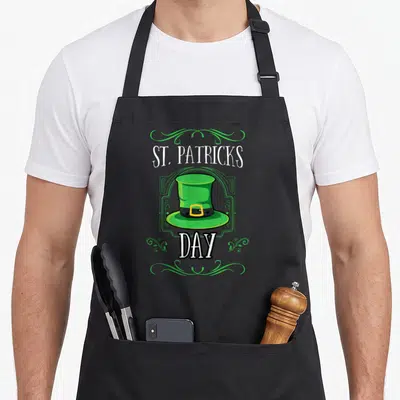 Tablier de cuisine noir avec chapeau vert irlandais et inscription St. Patricks Day entouré de motifs végétaux verts.