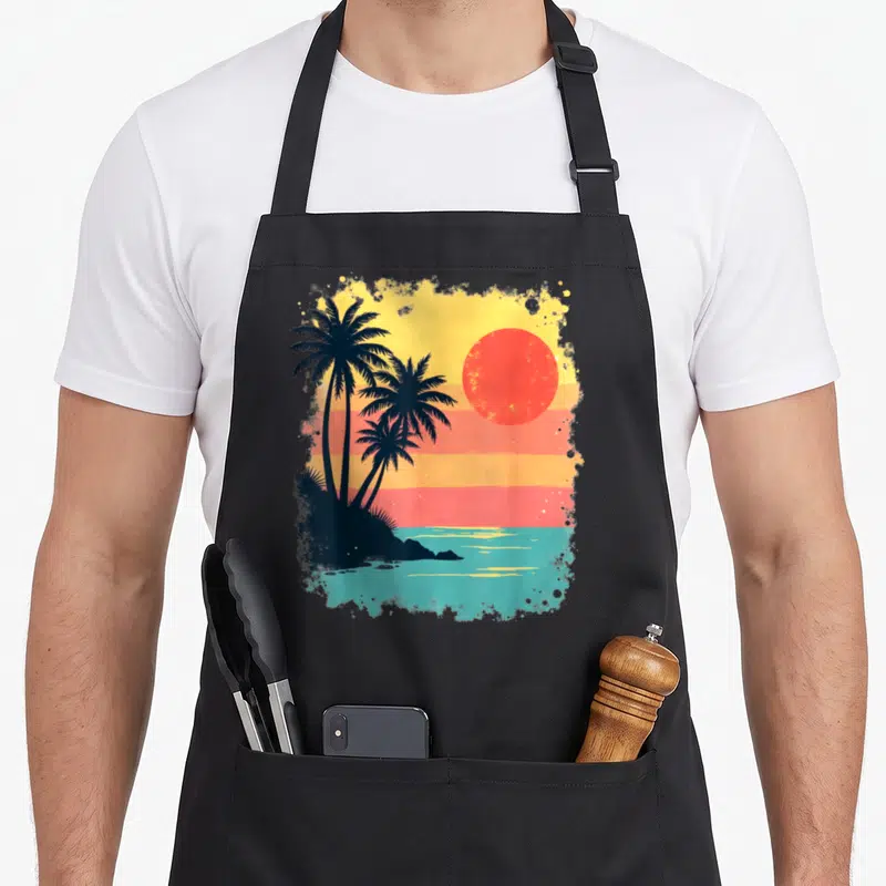 Tablier de cuisine noir avec illustration de palmiers et soleil couchant aux couleurs chaudes style rétro.