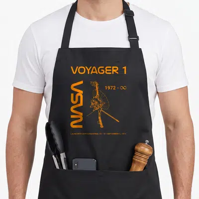 Tablier de cuisine noir avec illustration orange du vaisseau spatial Voyager 1 et texte NASA vertical.