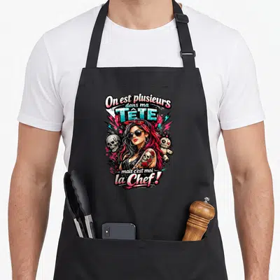 Tablier de cuisine noir avec une illustration rock d'une femme aux cheveux roses et lunettes de soleil, entourée de crânes et voodoo.