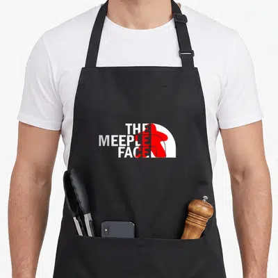 Tablier de cuisine noir avec une parodie du logo THE MEEPLE FACE en blanc et rouge sur le devant.