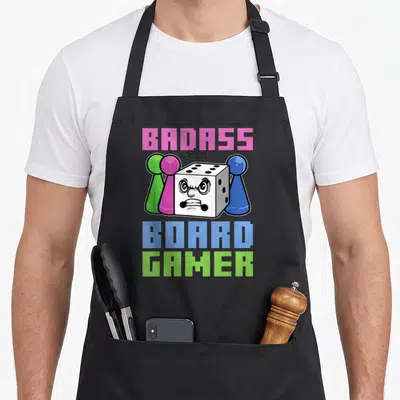 Tablier de cuisine noir avec un dé aux yeux furieux entouré de pions verts roses et bleus et du texte Badass Board Gamer.