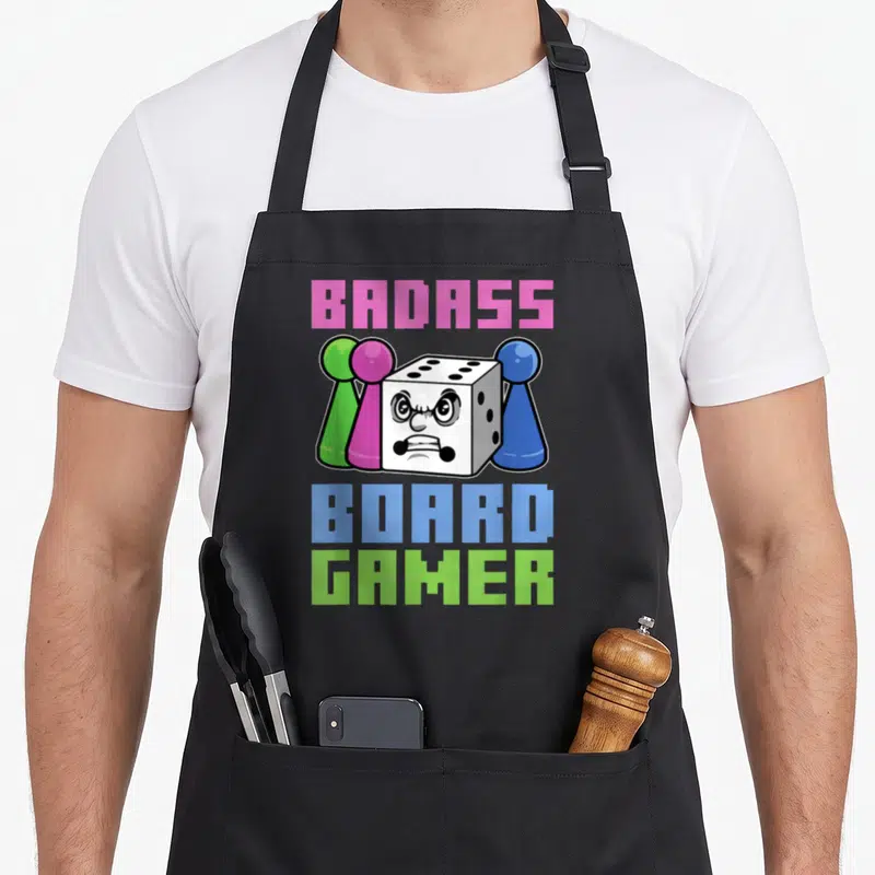 Tablier de cuisine noir avec un dé aux yeux furieux entouré de pions verts roses et bleus et du texte Badass Board Gamer.