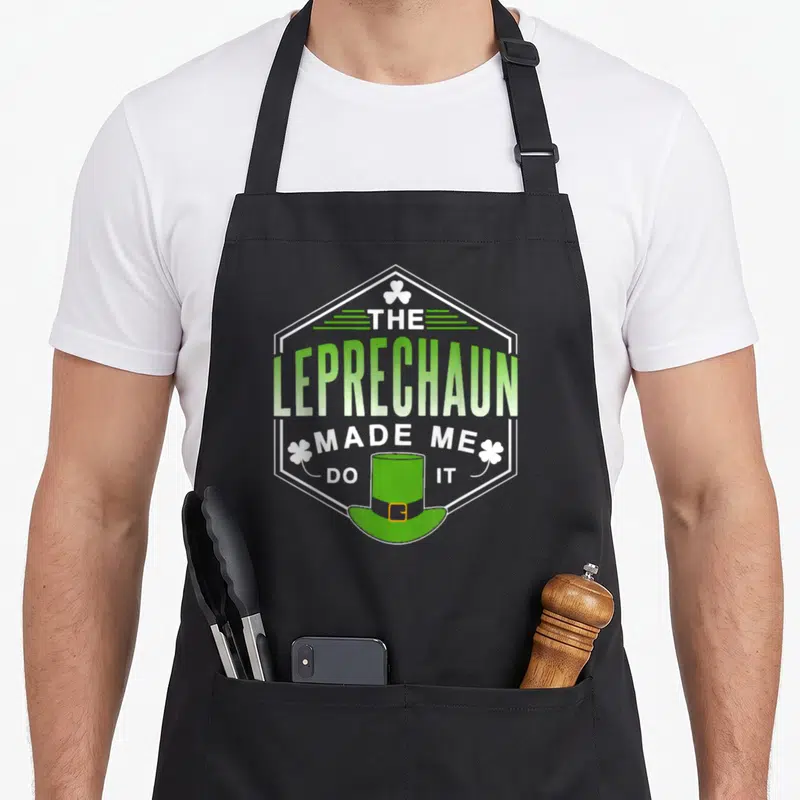 Tablier de cuisine noir avec un badge hexagonal vert et blanc affichant un chapeau de lutin et des trèfles à quatre feuilles.