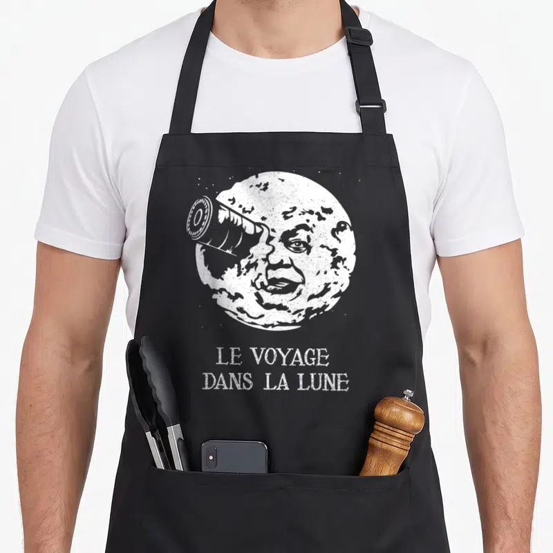 Tablier de cuisine noir avec une lune anthropomorphique regardant par un télescope et le titre du film imprimé en blanc.