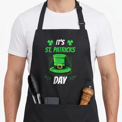 Tablier de cuisine noir avec un chapeau vert, des trèfles et les mots It's St. Patricks Day imprimés au centre.