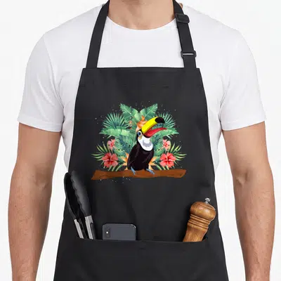 Tablier de cuisine noir avec un toucan coloré posé sur une branche au milieu de fleurs roses et de feuillage tropical vert.